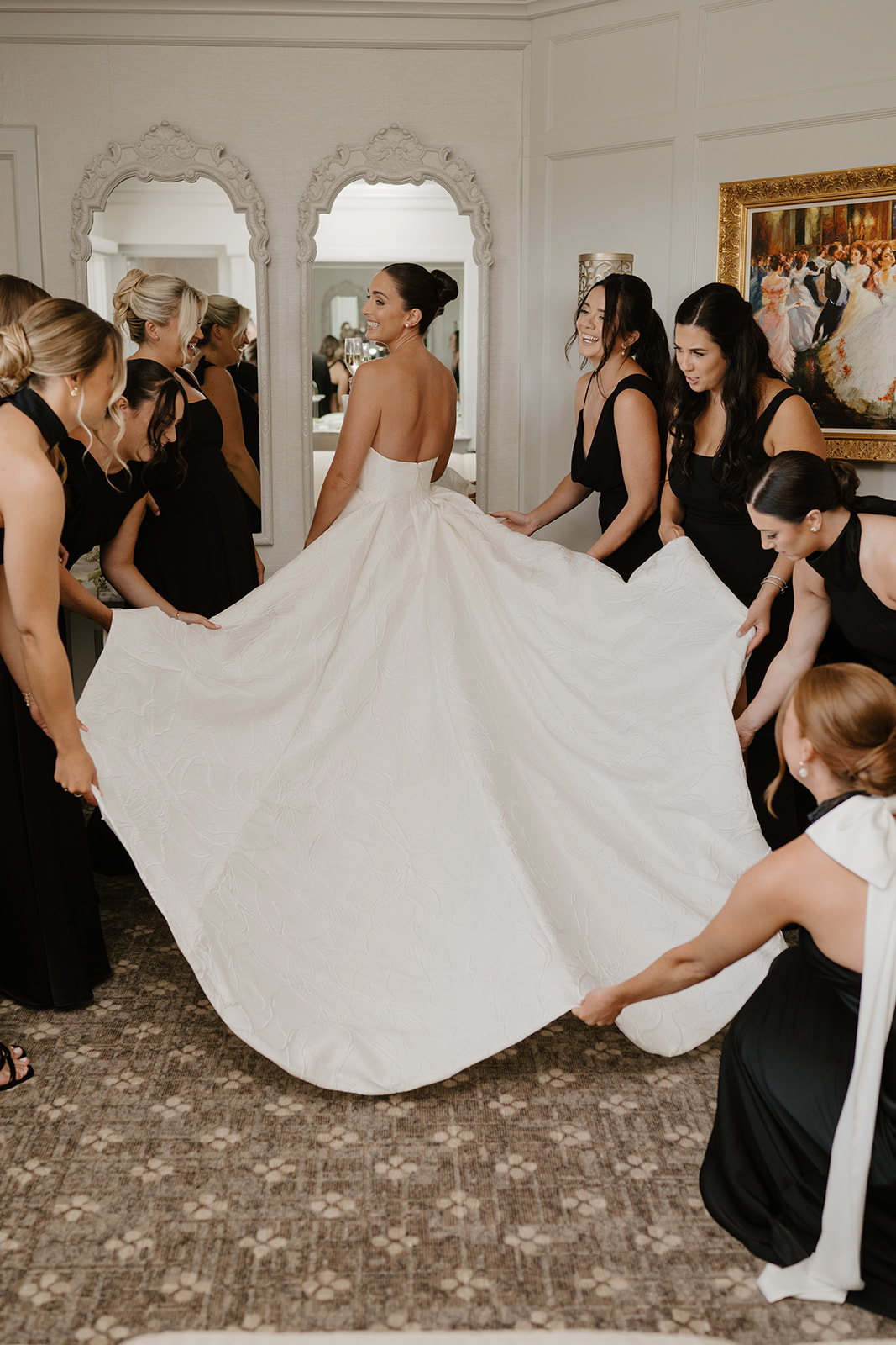 Bridesmaids holding up the full skirt of the bride's white gown as she smiles over her shoulder.